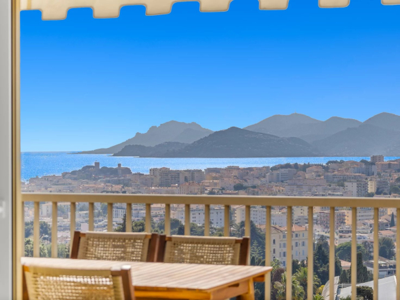 Outdoor balcony scene with a wooden table and woven chairs, railing in the foreground, overlooking a coastal town, sea, and distant blue mountains on a sunny day with a beige awning edge above.
