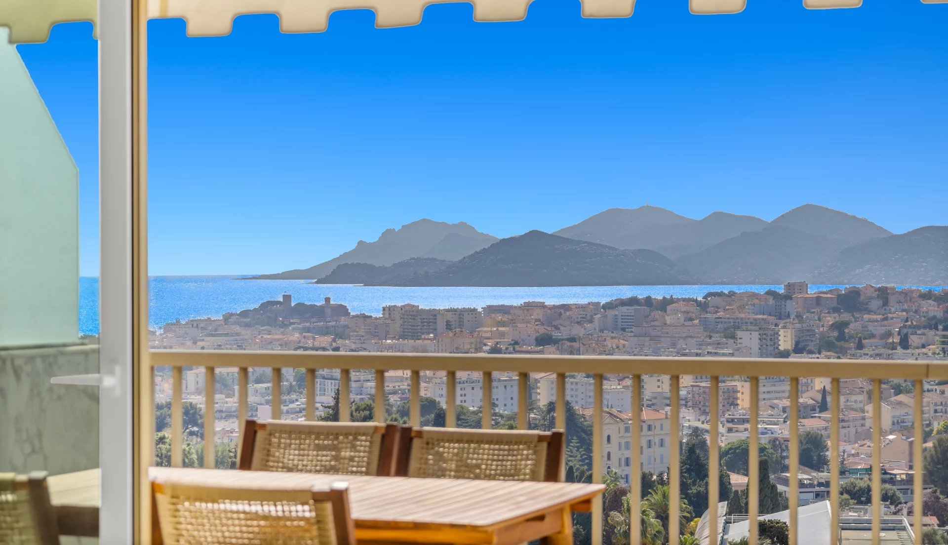 Outdoor balcony scene with a wooden table and woven chairs, railing in the foreground, overlooking a coastal town, sea, and distant blue mountains on a sunny day with a beige awning edge above.