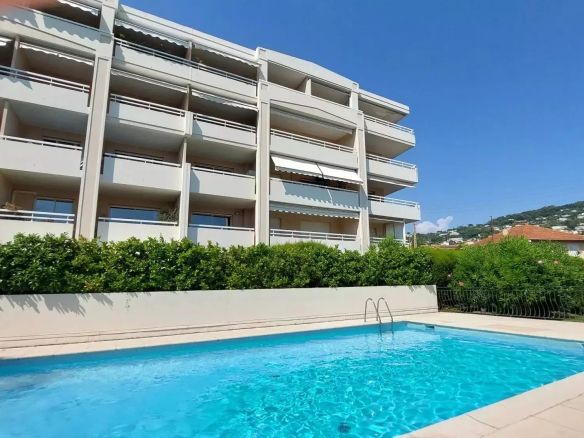 White multi-story apartment building with balconies and a blue swimming pool in the foreground under a clear blue sky.