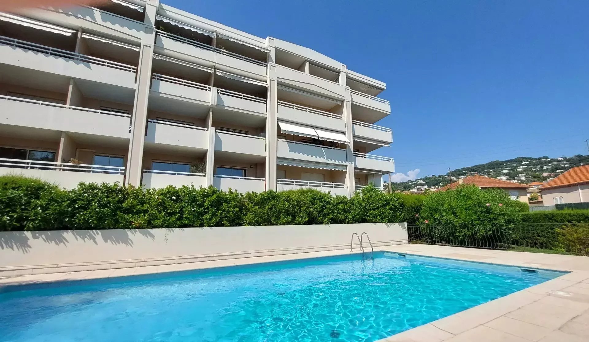 White multi-story apartment building with balconies and a blue swimming pool in the foreground under a clear blue sky.