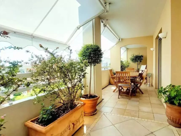 Sunlit balcony with wooden dining table and chairs, surrounded by potted plants in terracotta pots.
