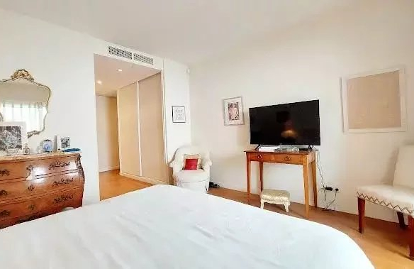 Bright bedroom with a white bed in the foreground, dresser and gold mirror on the left, and a TV on a wooden console across the room.