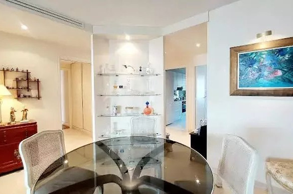 Bright dining area with a round glass table and white wicker chairs, glass display shelves in the center, a red sideboard with a lamp to the left, and a blue-framed painting on the right; an open kitchen doorway visible in the back.