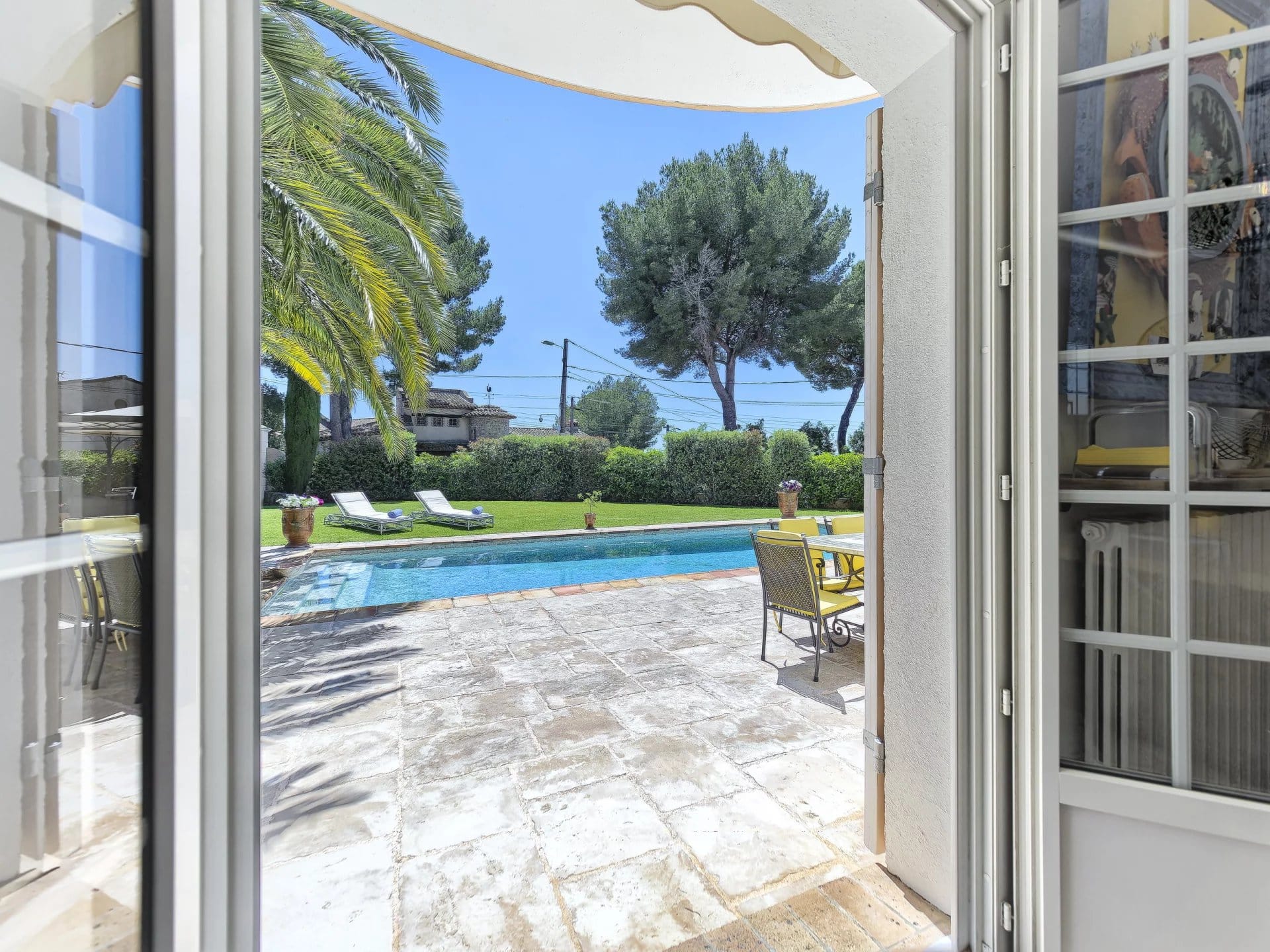 View of a swimming pool and garden from inside a bastide in Le Cannet, with palm trees and sun loungers.