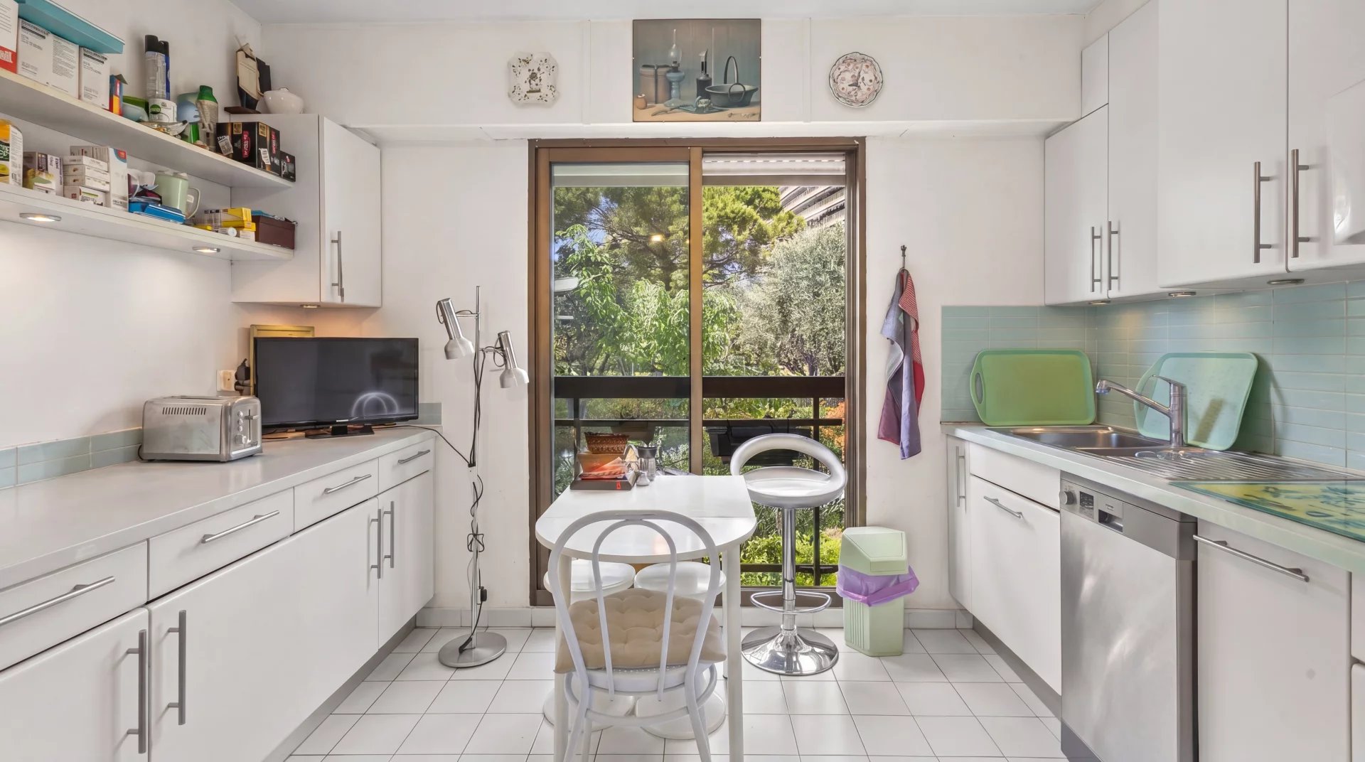 Cuisine lumineuse avec vue sur un jardin, équipée de meubles blancs et d'une table pour deux.