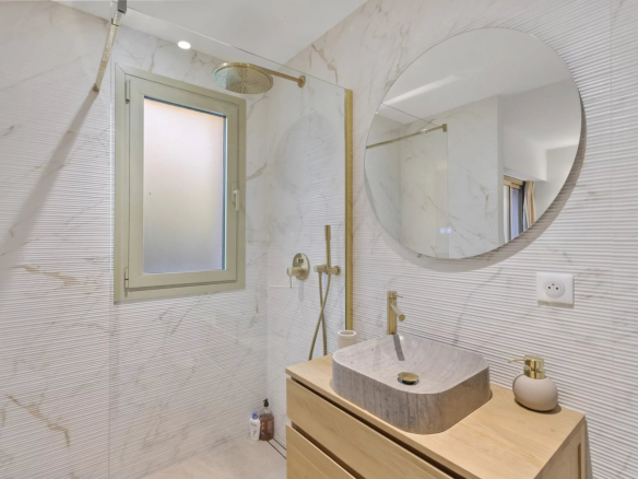 Modern bathroom with shower and wooden furniture, marble tiles, window and round mirror.