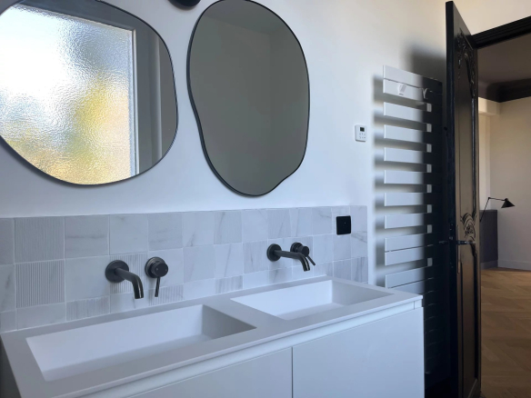 Salle de bain moderne avec double vasque et miroirs ovales, murs en carrelage blanc, et radiateur design.