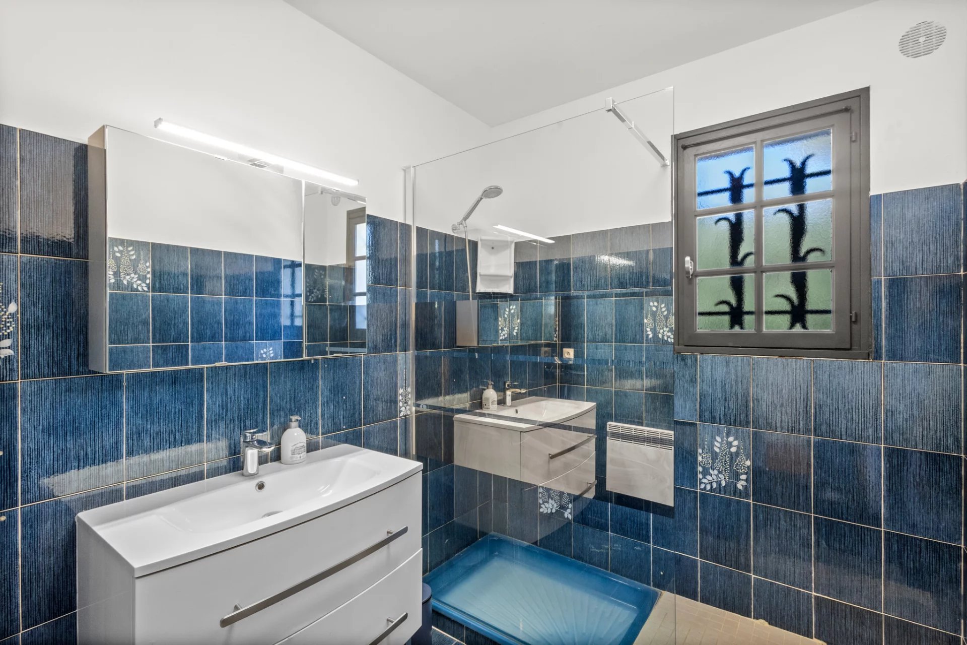 Modern bathroom with blue tiles and vanity unit, window with patterns.