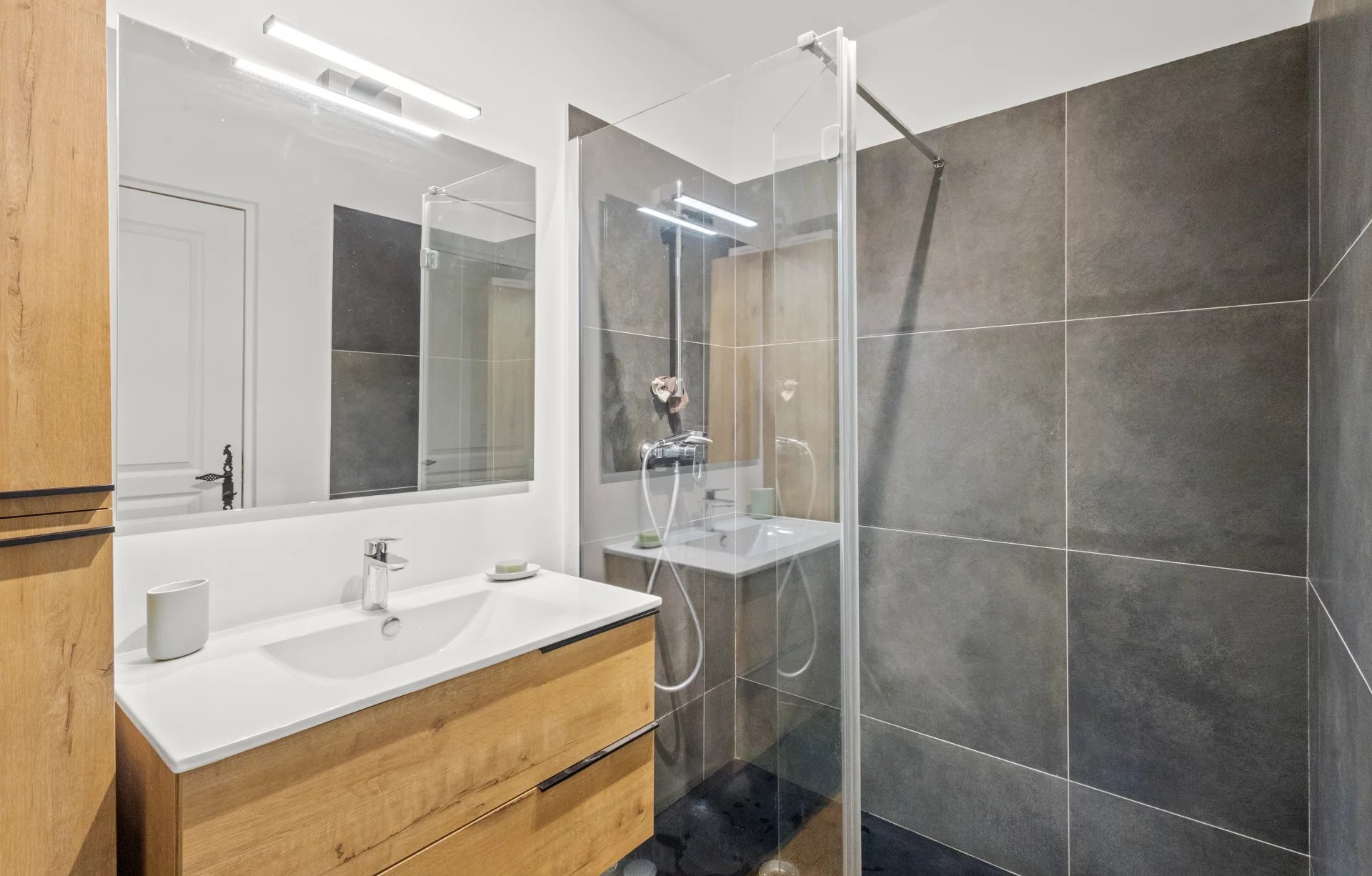 Salle de bain moderne avec douche, meuble en bois et miroir, carrelage gris.