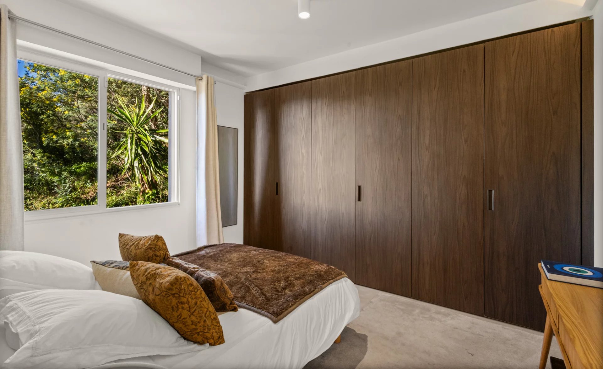 Chambre d’appartement rénové à Cannes Chambre d'un appartement rénové avec lit et placard en bois, vue sur des plantes à l'extérieur.