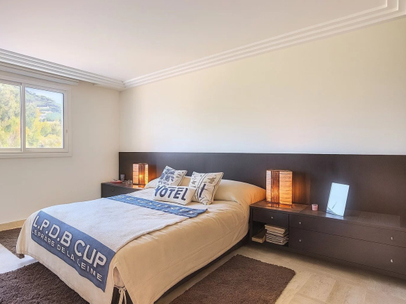 Bright hotel-style bedroom with a low dark wood headboard, two matching nightstands, and warm table lamps flanking the bed; large window shows greenery outside; beige bedding with a blue accent blanket.