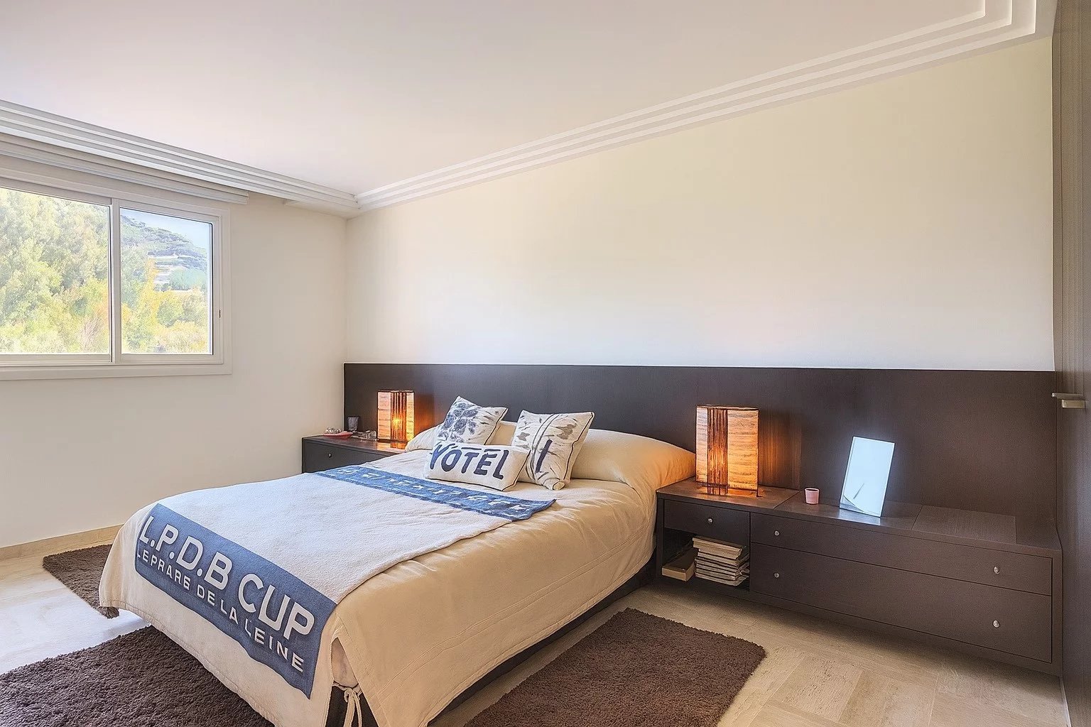 Bright hotel-style bedroom with a low dark wood headboard, two matching nightstands, and warm table lamps flanking the bed; large window shows greenery outside; beige bedding with a blue accent blanket.