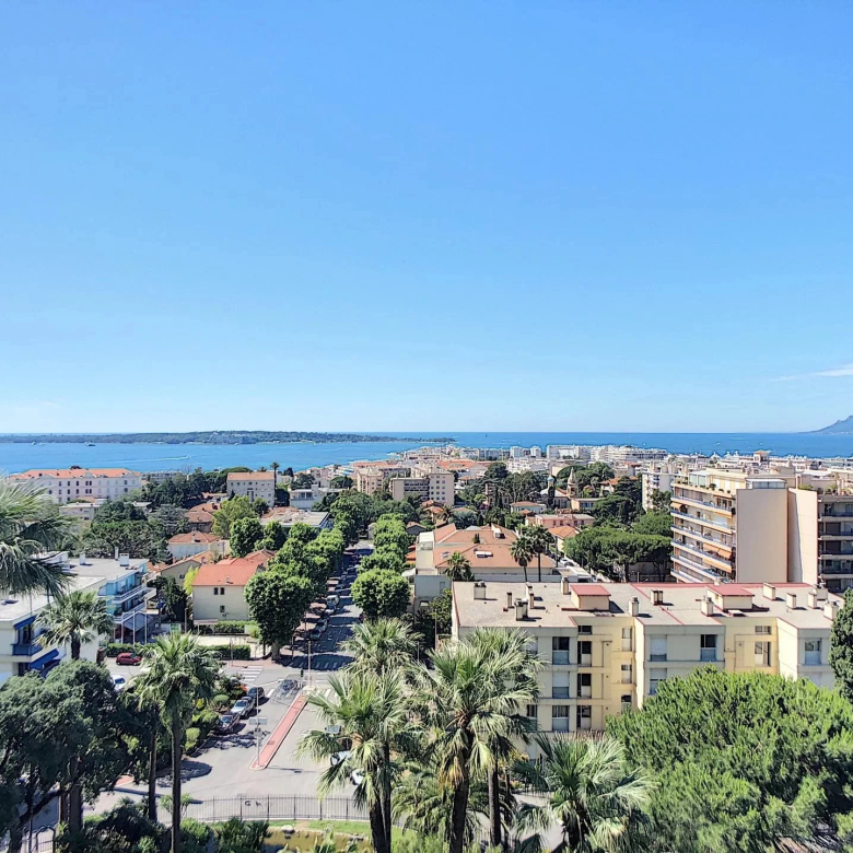Aerial view of a coastal city with palm trees, orange-tiled roofs, and the blue sea on a sunny day.