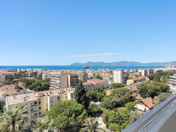 Wide coastal cityscape viewed from a balcony: buildings, greenery, and palm trees with the sea and distant mountains under a clear blue sky.