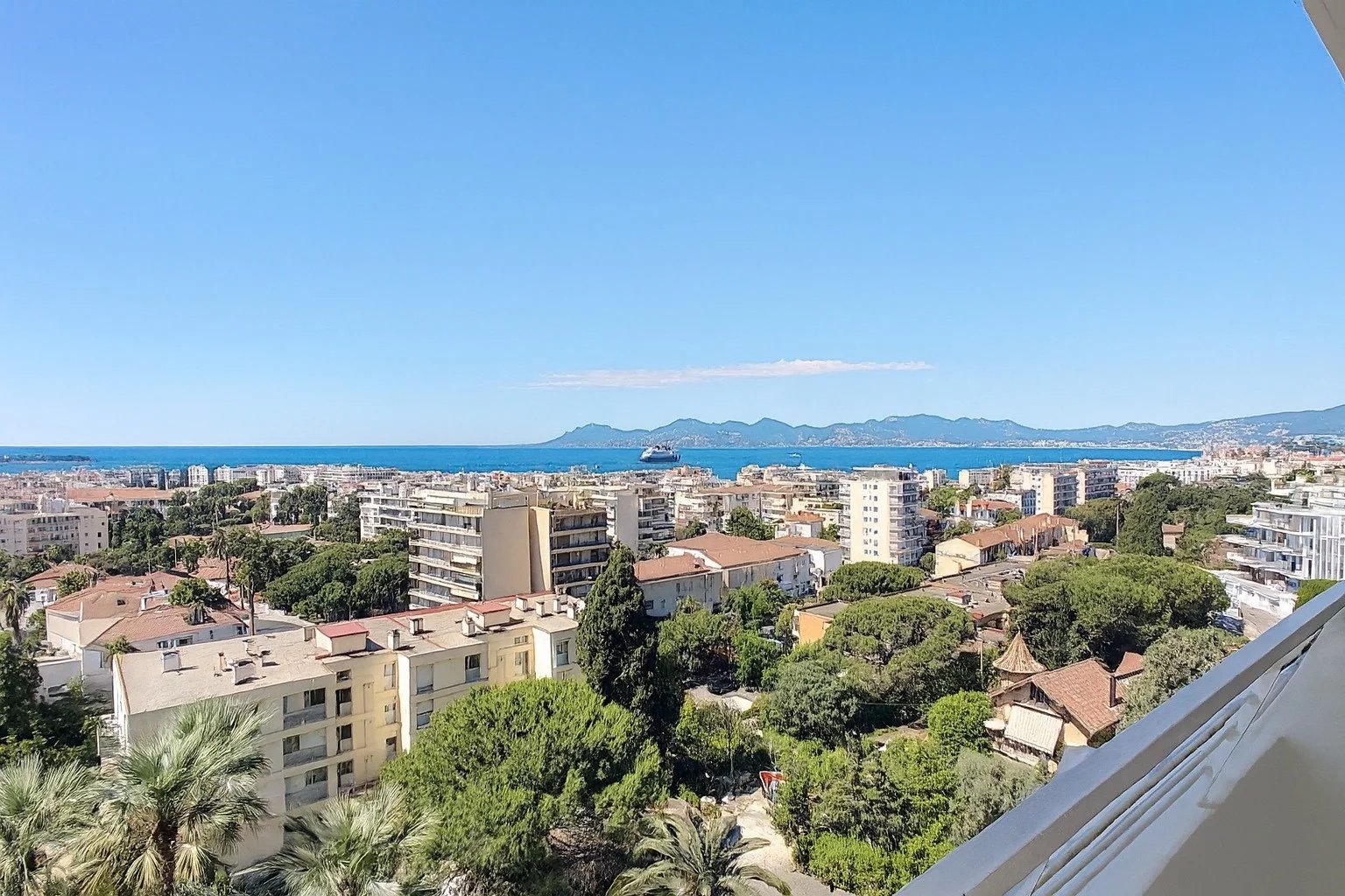 Wide coastal cityscape viewed from a balcony: buildings, greenery, and palm trees with the sea and distant mountains under a clear blue sky.