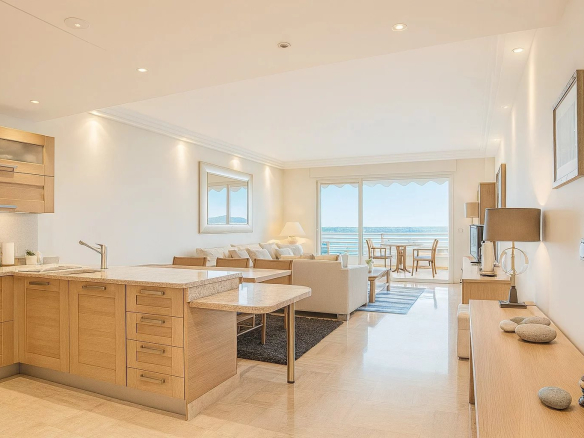 Bright open-plan living area with light wood kitchen cabinets, beige furniture, and a seaside balcony view through sliding doors.