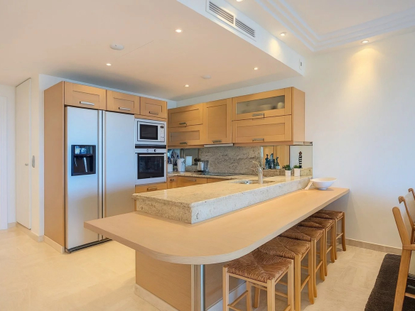 Bright kitchen with light wood cabinets, stainless steel fridge, and a long granite island with seating for five.