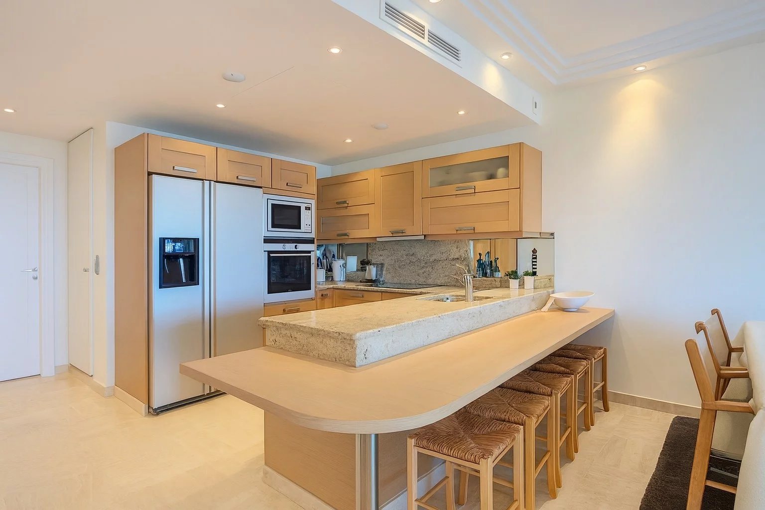 Bright kitchen with light wood cabinets, stainless steel fridge, and a long granite island with seating for five.