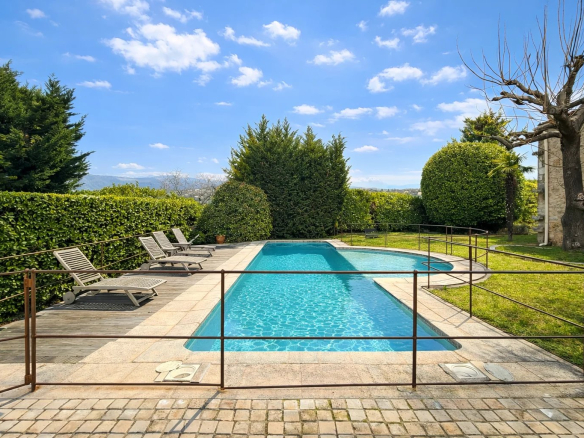 Swimming pool surrounded by sun loungers in a green garden with hedges.