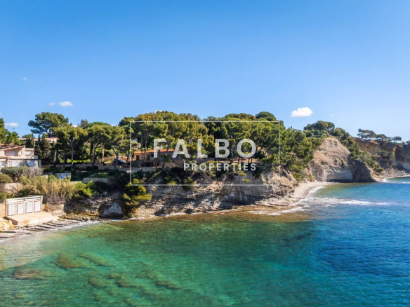 Coastal scene with rocky cliff, pine trees, and a turquoise sea along a shoreline with houses in the background.