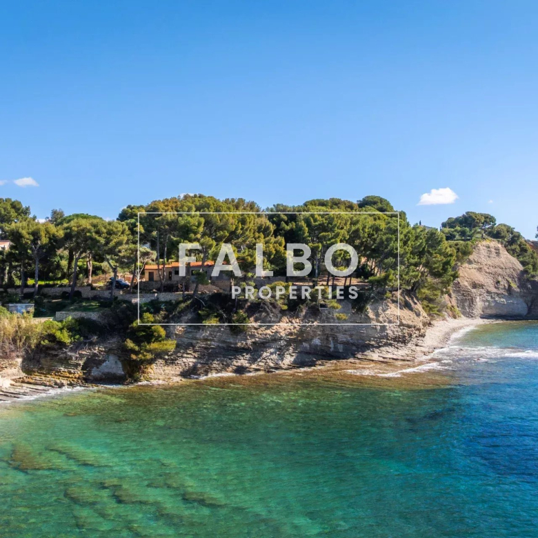 Coastal scene with rocky cliff, pine trees, and a turquoise sea along a shoreline with houses in the background.