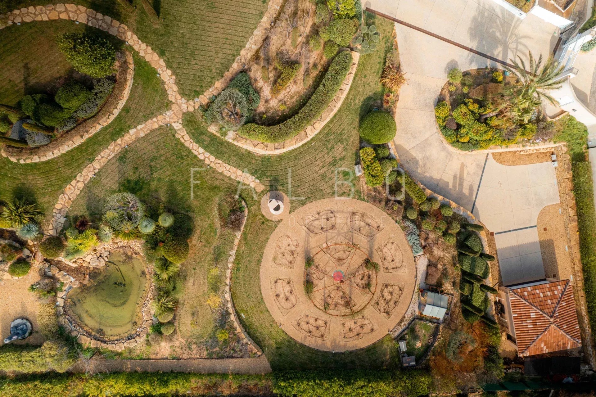 Vue aérienne d'un parc paysager avec cheminements et aménagements, villa en arrière-plan.