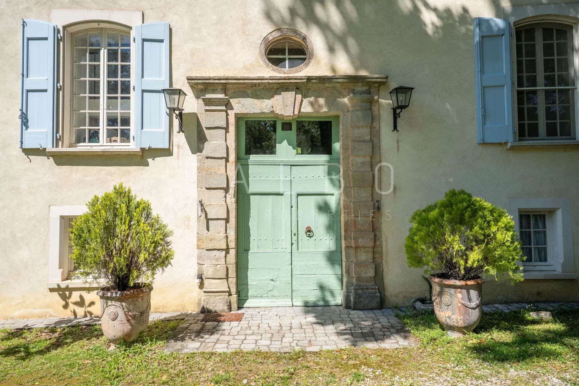 Entrée d’un château restauré près d’Anduze Entrée d'un château restauré avec porte verte et fenêtres à volets bleus, entourée de plantes en pot.