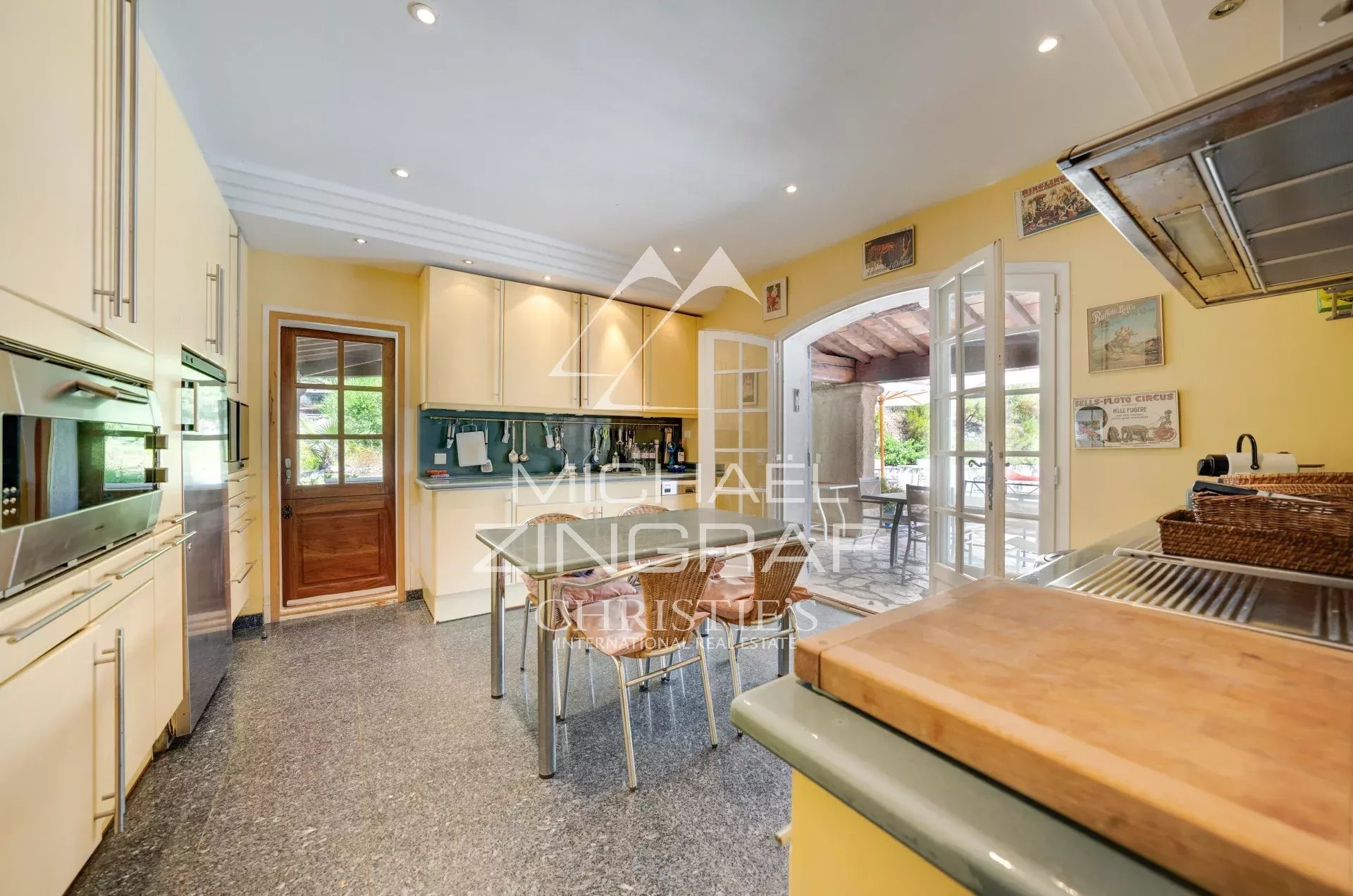Bright kitchen with yellow walls, white cabinets, stainless appliances, a small table and wicker chairs, and French doors to an outdoor patio.