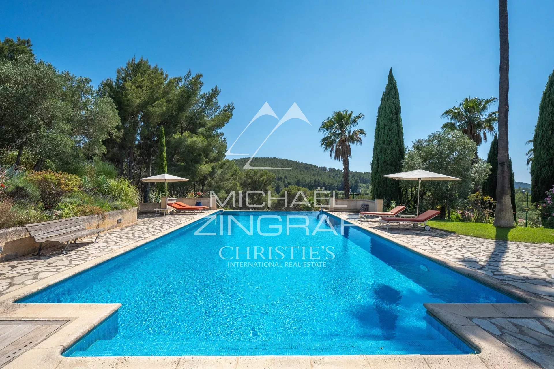 Luxurious rectangular pool with stone deck, lounge chairs and umbrellas under a bright blue sky.