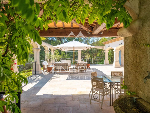 Sunny stone terrace with arched columns, a white umbrella over a seating area, and lush greenery framing the scene.