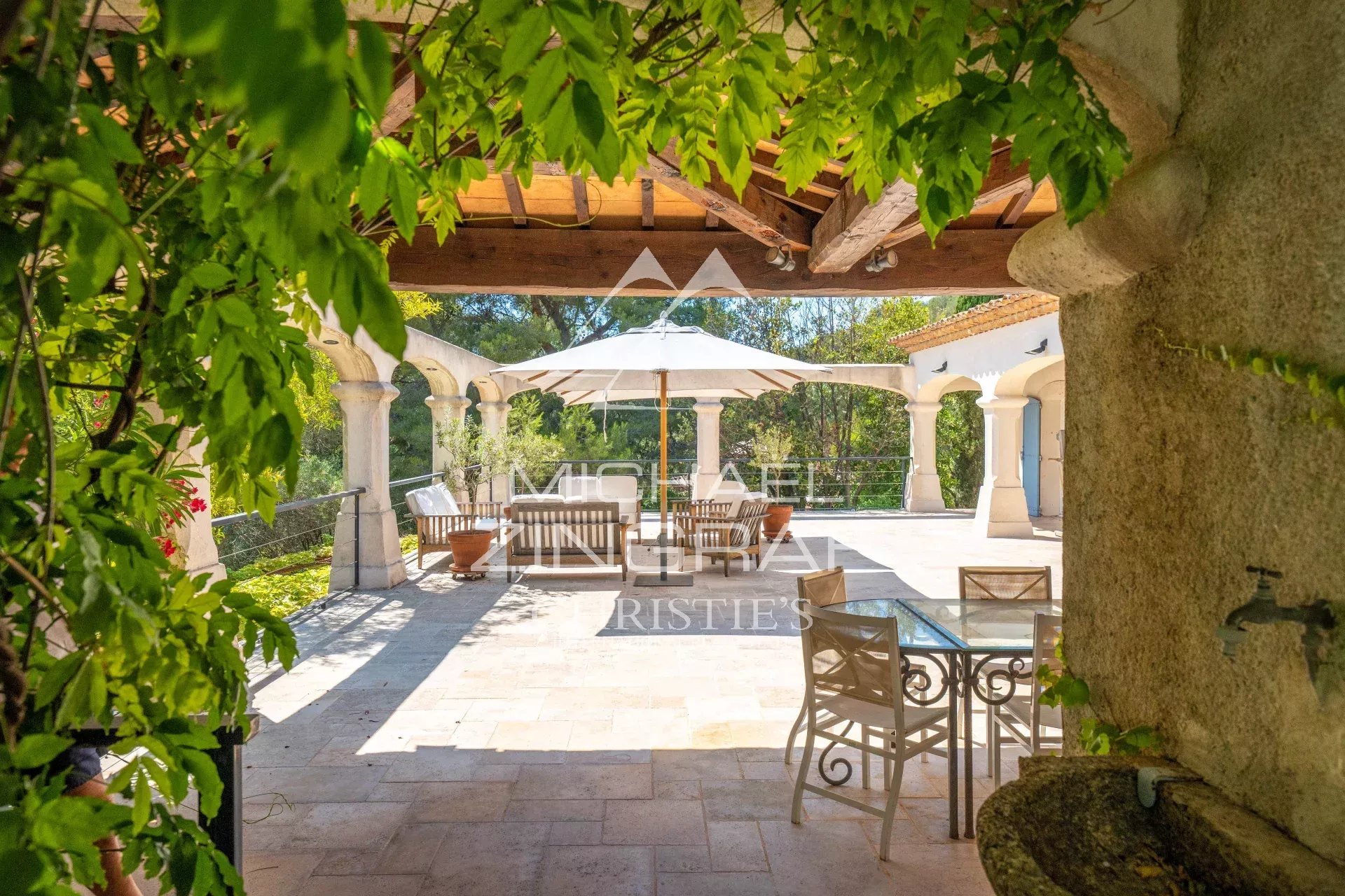 Sunny stone terrace with arched columns, a white umbrella over a seating area, and lush greenery framing the scene.