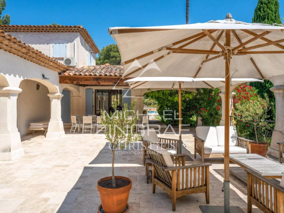 Sunny Mediterranean courtyard with beige stone floor, white arches, and wooden seating under large umbrellas in bright daylight.