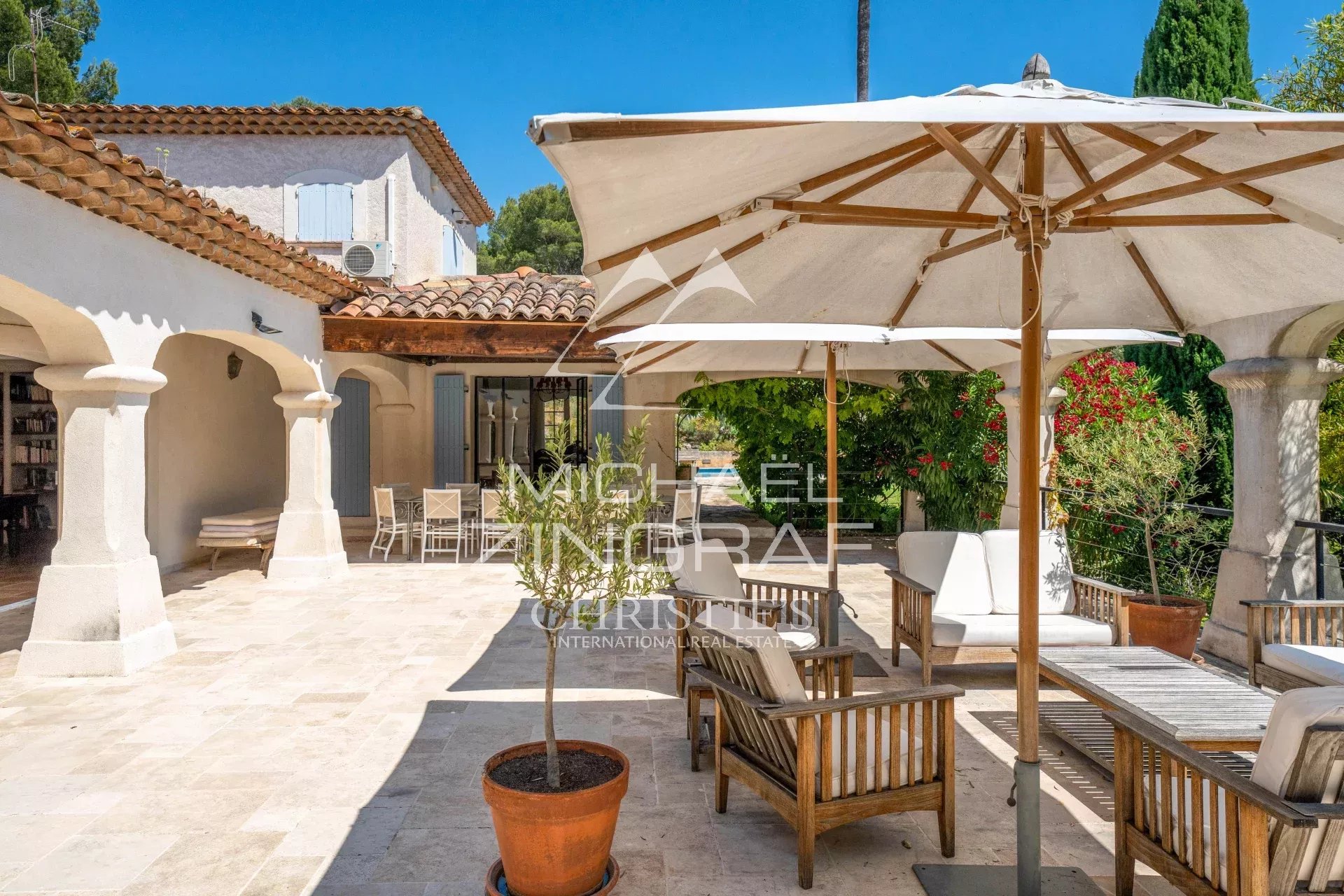 Sunny Mediterranean courtyard with beige stone floor, white arches, and wooden seating under large umbrellas in bright daylight.