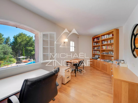 Bright home office with a large arched window, a wooden desk, and a tall bookcase overlooking a pool and garden outside