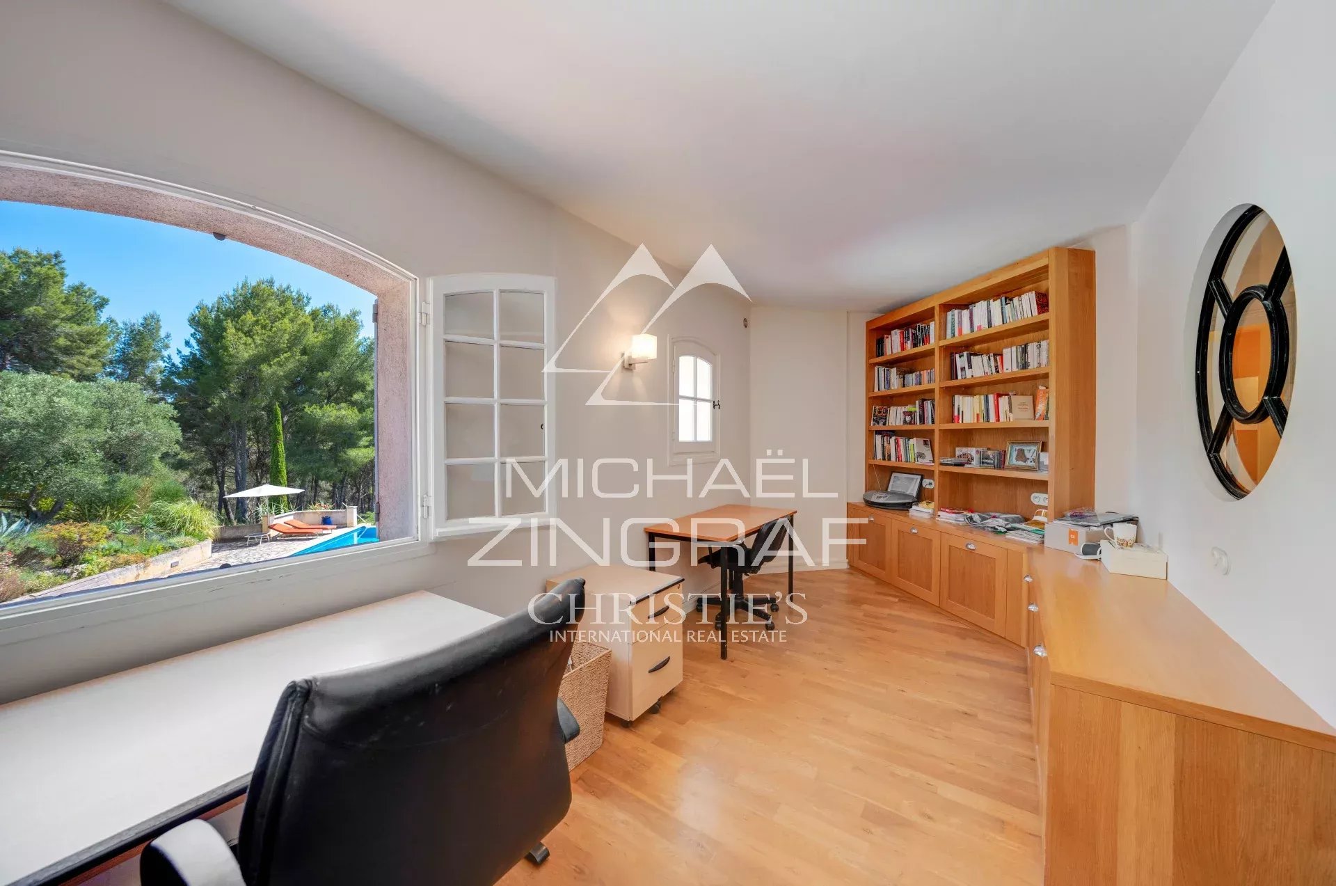 Bright home office with a large arched window, a wooden desk, and a tall bookcase overlooking a pool and garden outside