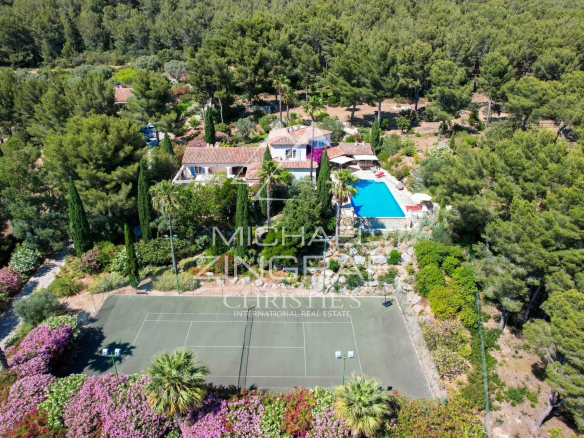 Aerial view of a villa with a blue swimming pool, surrounding trees, and a tennis court at the edge of a landscaped garden.