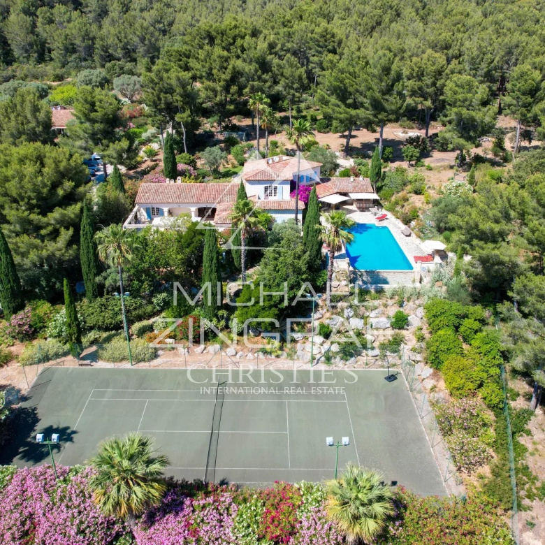 Aerial view of a villa with a blue swimming pool, surrounding trees, and a tennis court at the edge of a landscaped garden.