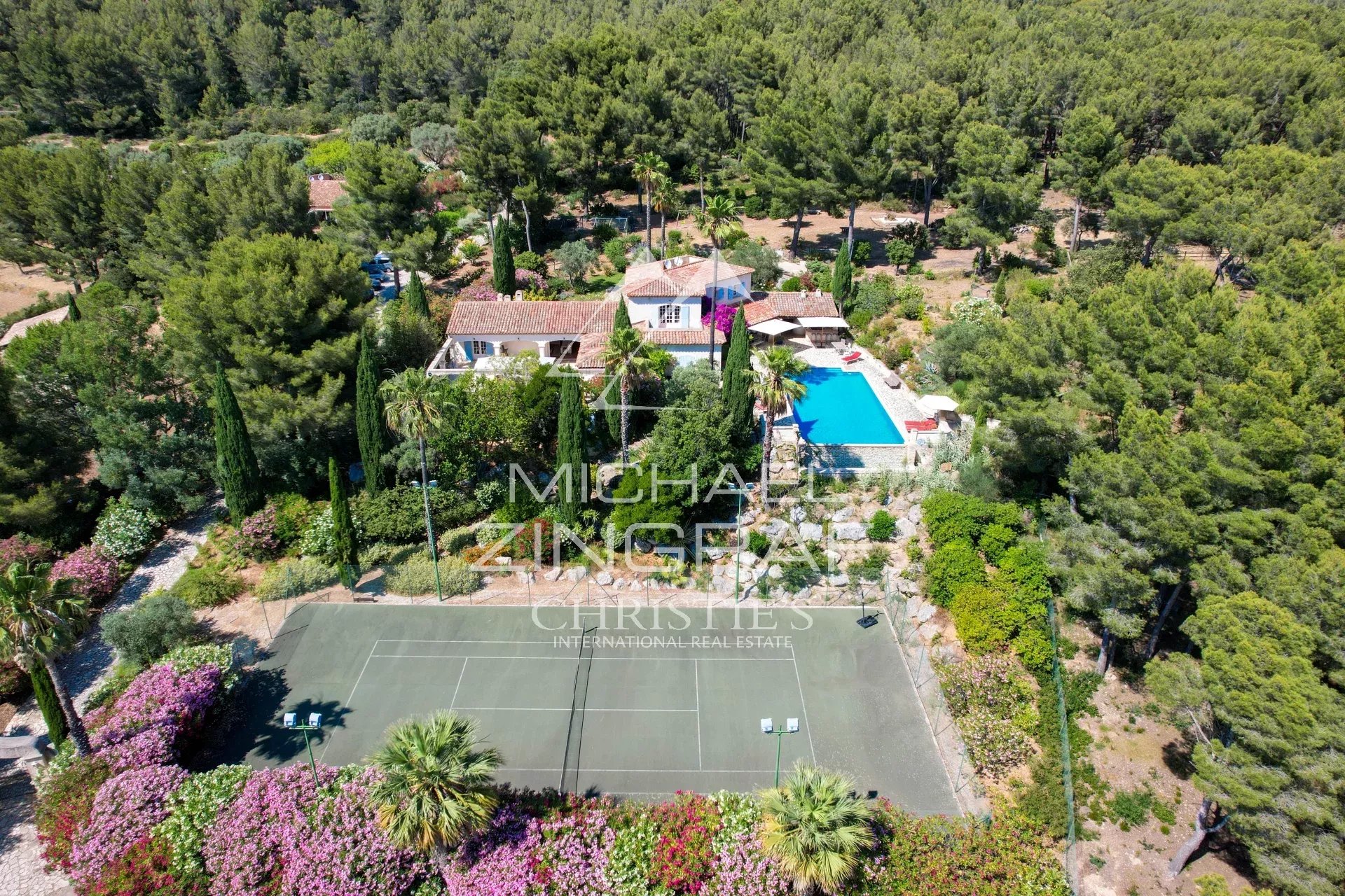 Aerial view of a villa with a blue swimming pool, surrounding trees, and a tennis court at the edge of a landscaped garden.