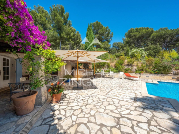 Sunny outdoor pool patio with stone flooring, white umbrellas, lounge chairs, and vibrant purple bougainvillea against a blue sky.