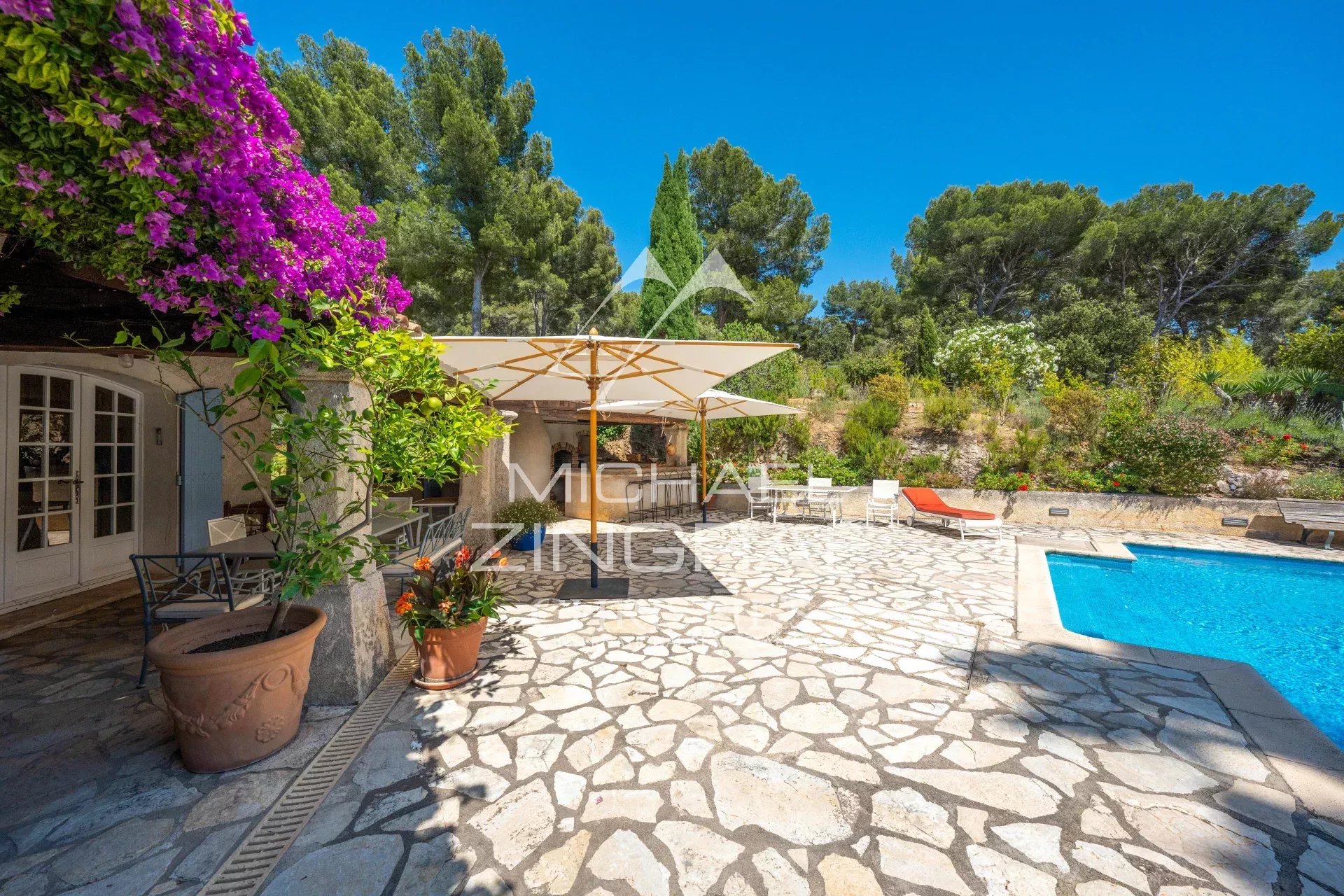 Sunny outdoor pool patio with stone flooring, white umbrellas, lounge chairs, and vibrant purple bougainvillea against a blue sky.