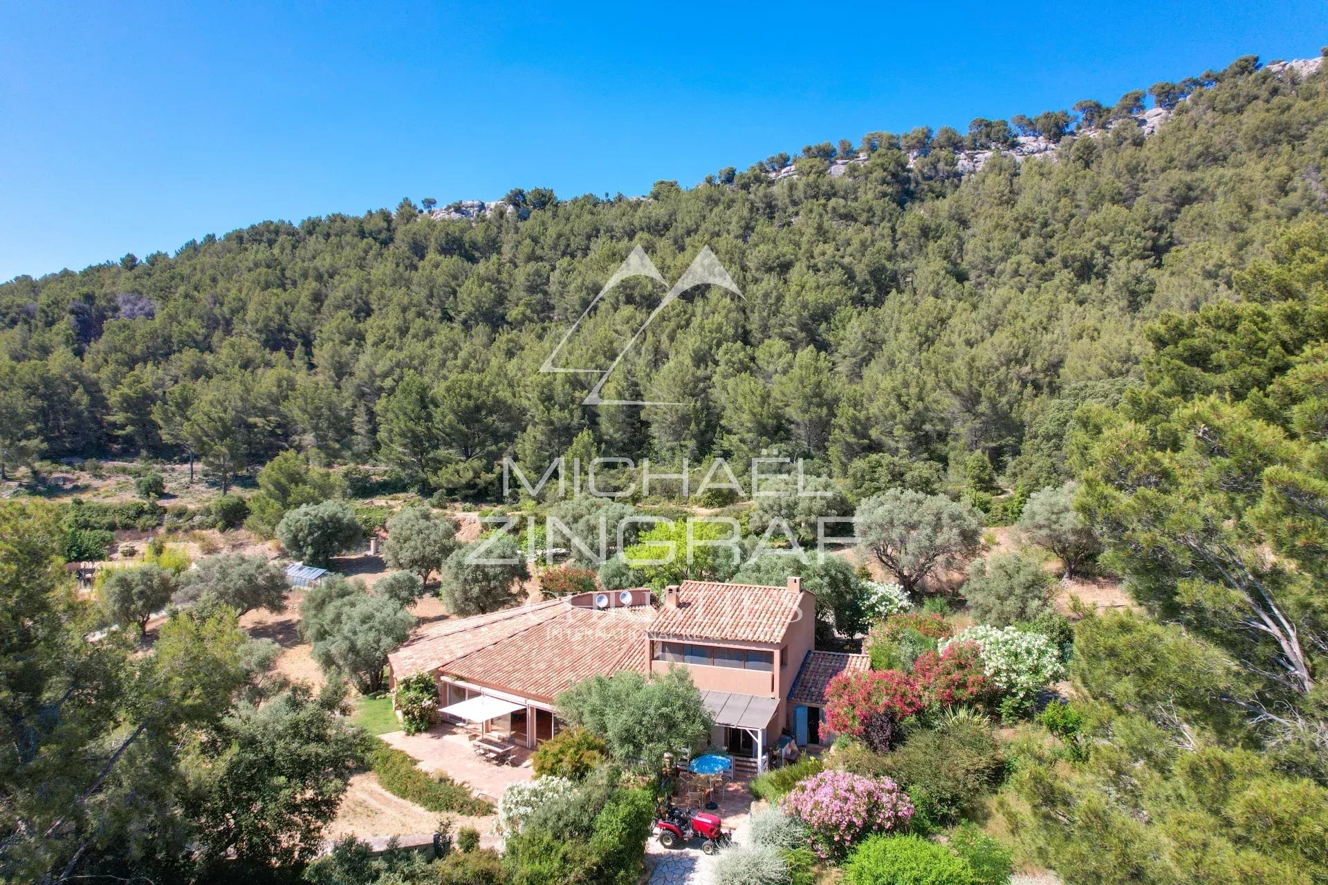 Drone view of a house with orange-tiled roof nestled among trees on a hillside, bright blue sky overhead, with a lush garden surrounding the property.