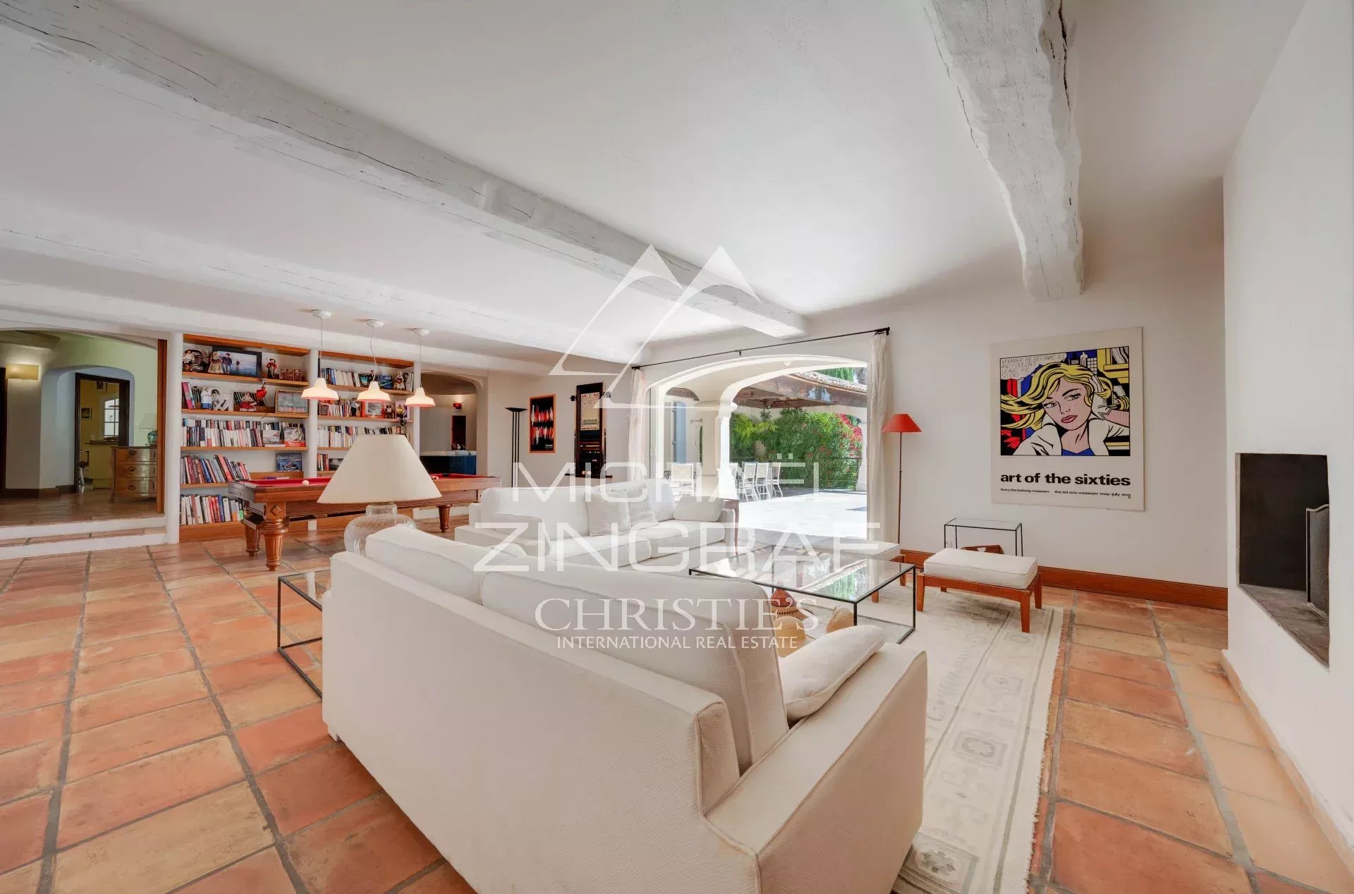 Bright living room with white sofas, glass coffee table, terracotta tile floor, and a bookshelf wall in the background.