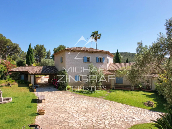 Mediterranean-style villa with a terracotta tile roof, blue-shuttered windows, and a stone driveway surrounded by a green garden.