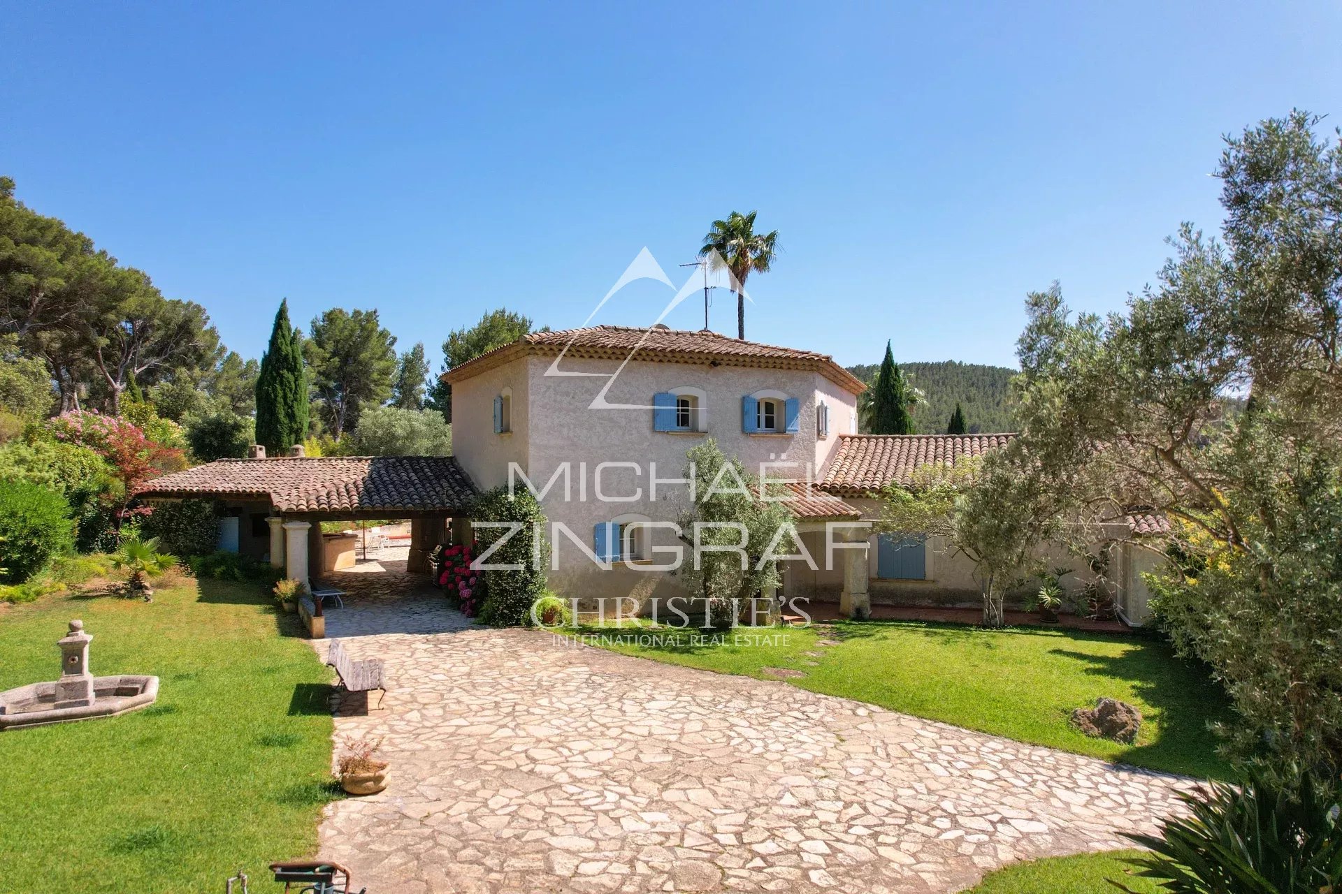 Mediterranean-style villa with a terracotta tile roof, blue-shuttered windows, and a stone driveway surrounded by a green garden.