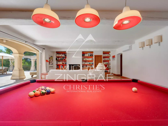 Bright game room with a red billiard/pool table, balls racked on the table, white seating and bookshelves in the background, and large open doors revealing an outdoor patio with umbrellas. Natural light fills the space, and a watermark overlaps the center of the image.