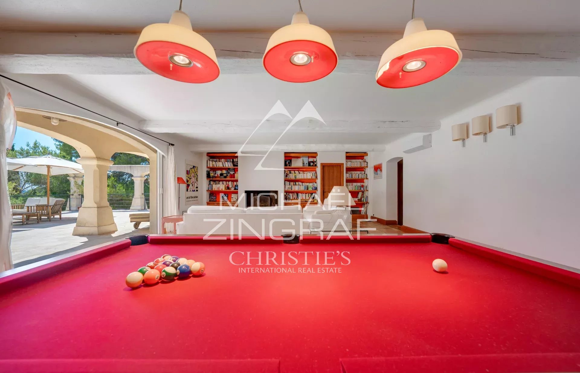 Bright game room with a red billiard/pool table, balls racked on the table, white seating and bookshelves in the background, and large open doors revealing an outdoor patio with umbrellas. Natural light fills the space, and a watermark overlaps the center of the image.