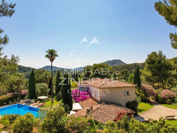 Mediterranean villa with a terracotta roof beside a blue swimming pool, palm trees and a lush garden under a clear sky.