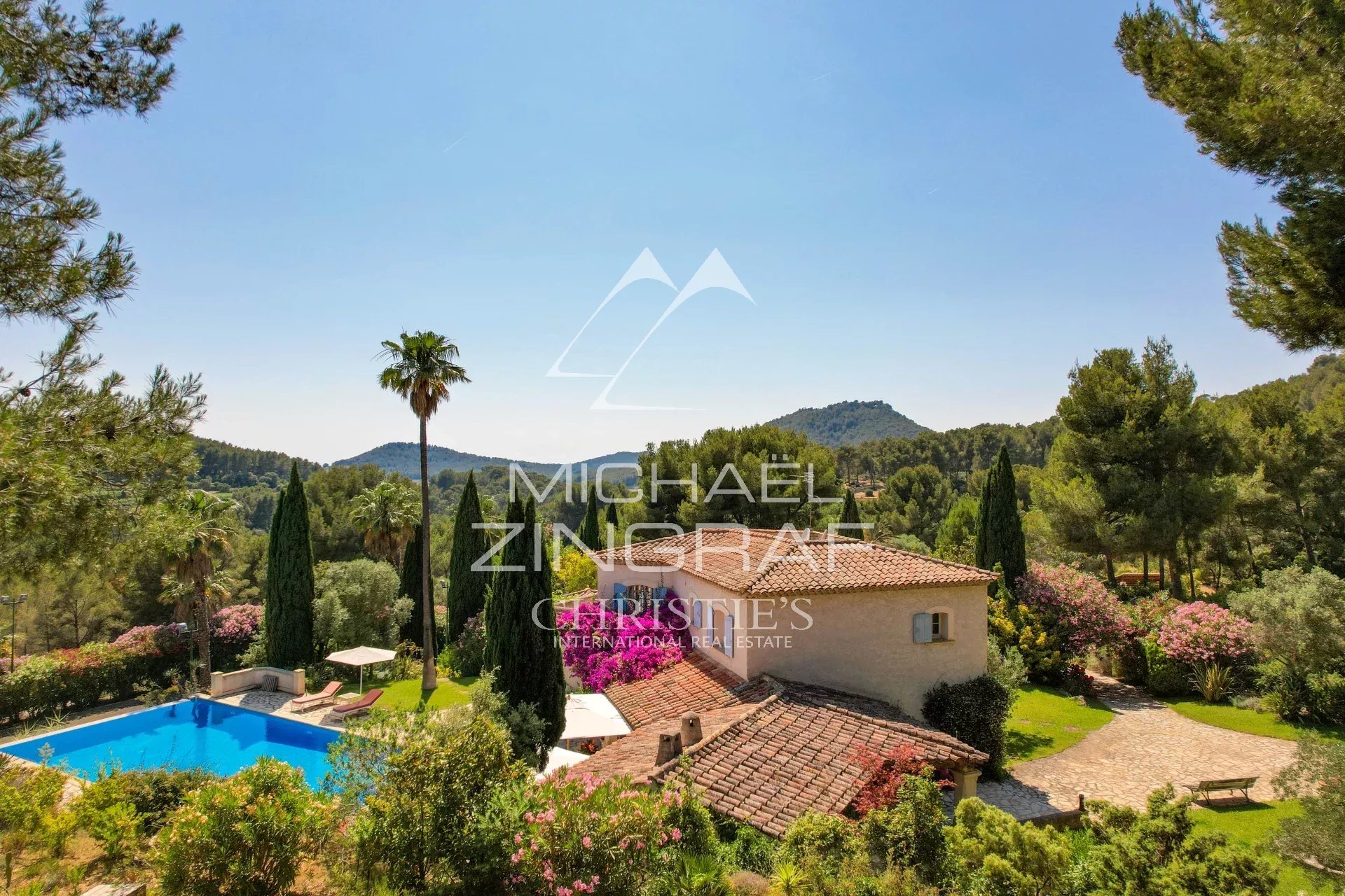 Mediterranean villa with a terracotta roof beside a blue swimming pool, palm trees and a lush garden under a clear sky.