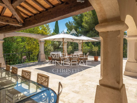 Stone-arched terrace with a wooden beam ceiling, an outdoor glass dining table and chairs, and a large white umbrella over a seating area overlooking greenery
