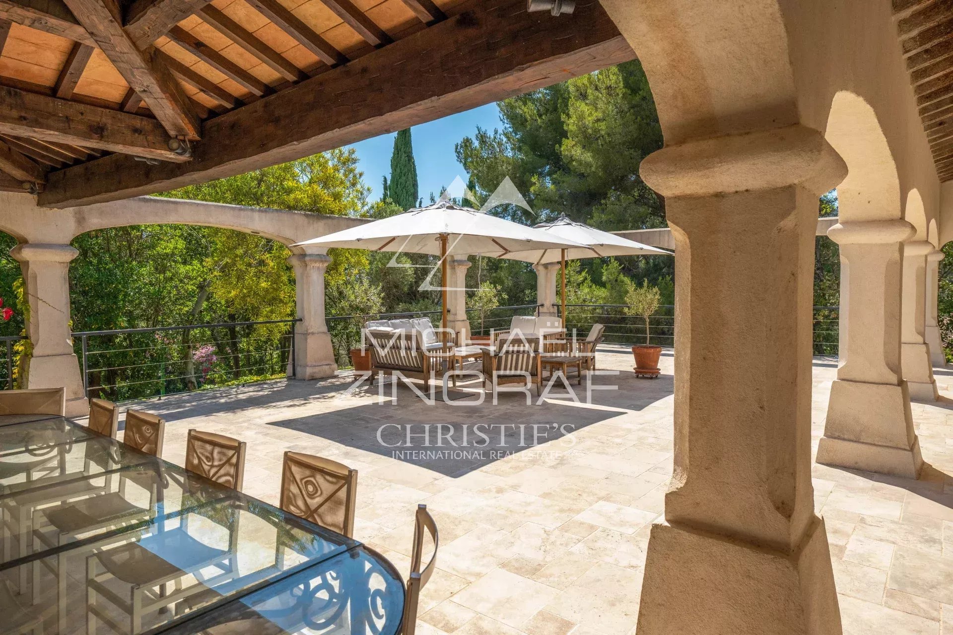 Stone-arched terrace with a wooden beam ceiling, an outdoor glass dining table and chairs, and a large white umbrella over a seating area overlooking greenery